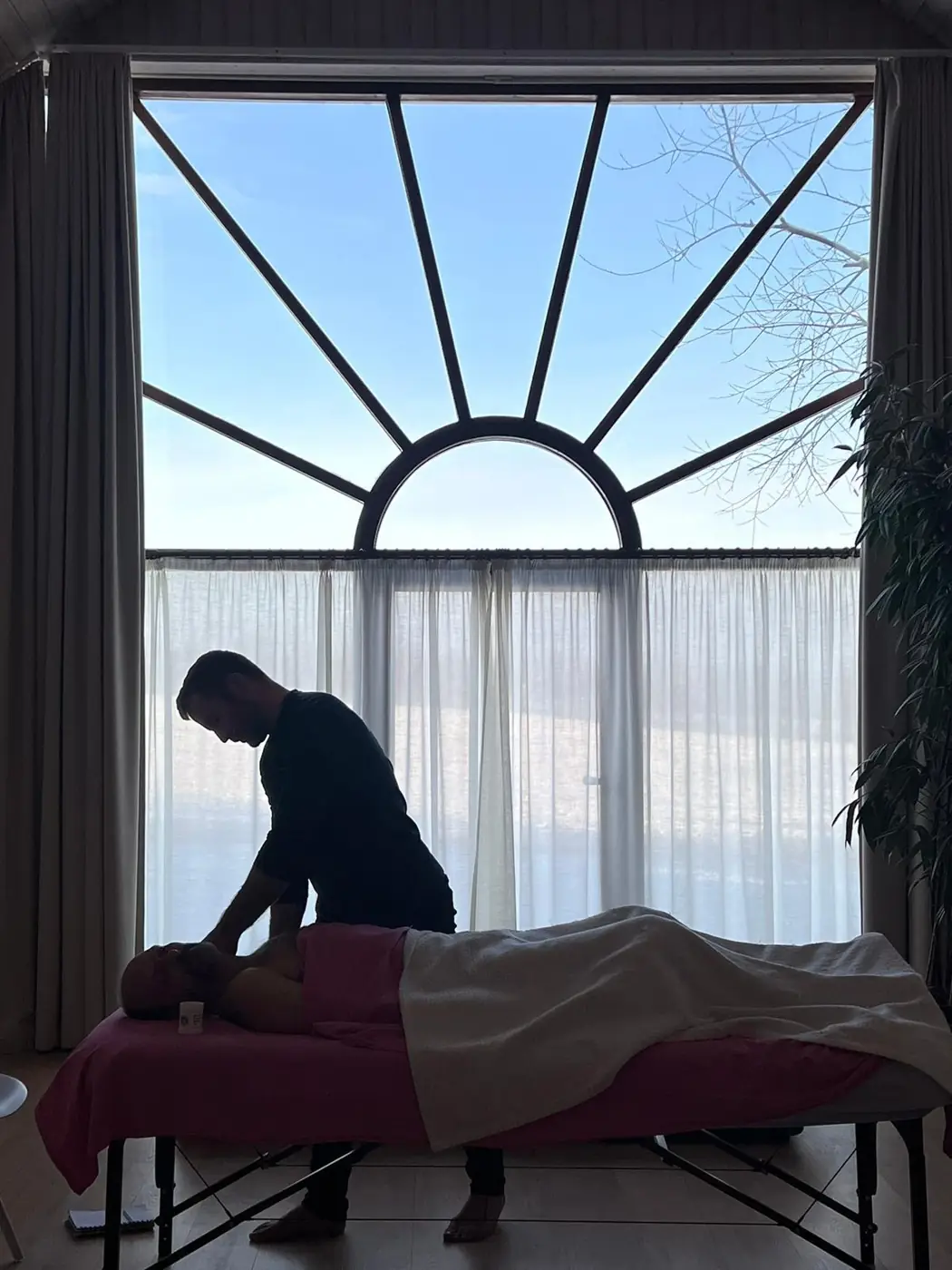 Silhouette of Pat working with a client on a massage table in front of a large sunburst window