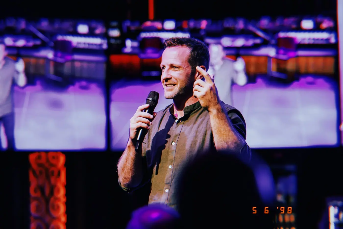 Pat speaking on stage with a microphone in front of large screens